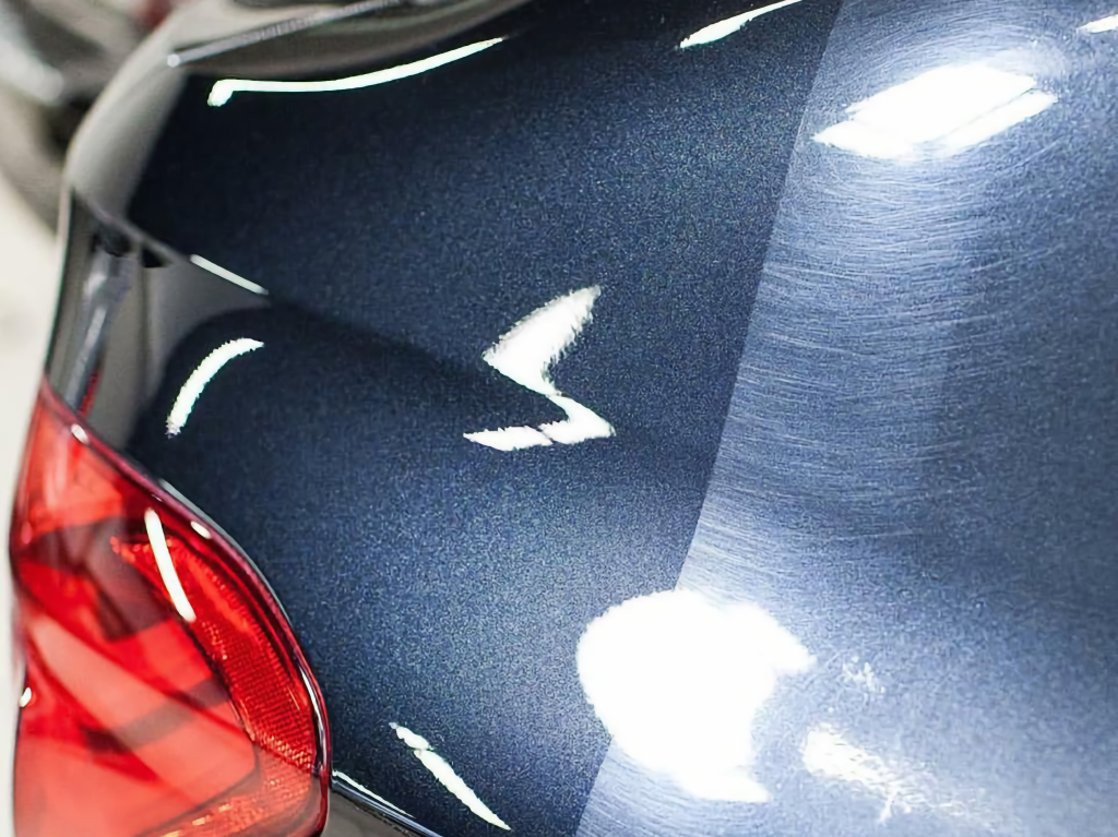 Close-up of a dark blue car panel with white scratches and a red tail light visible at the left edge.