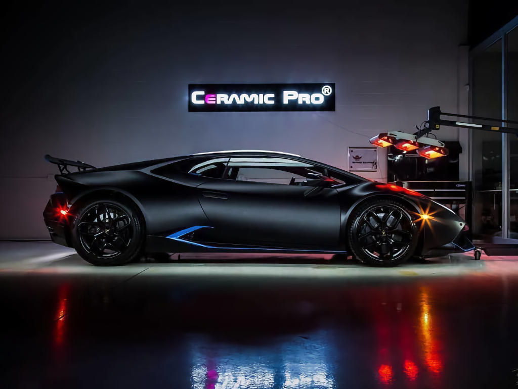 Matte black exotic sports car in a showroom beneath a Ceramic Pro sign, with red taillights and reflected floor lights.