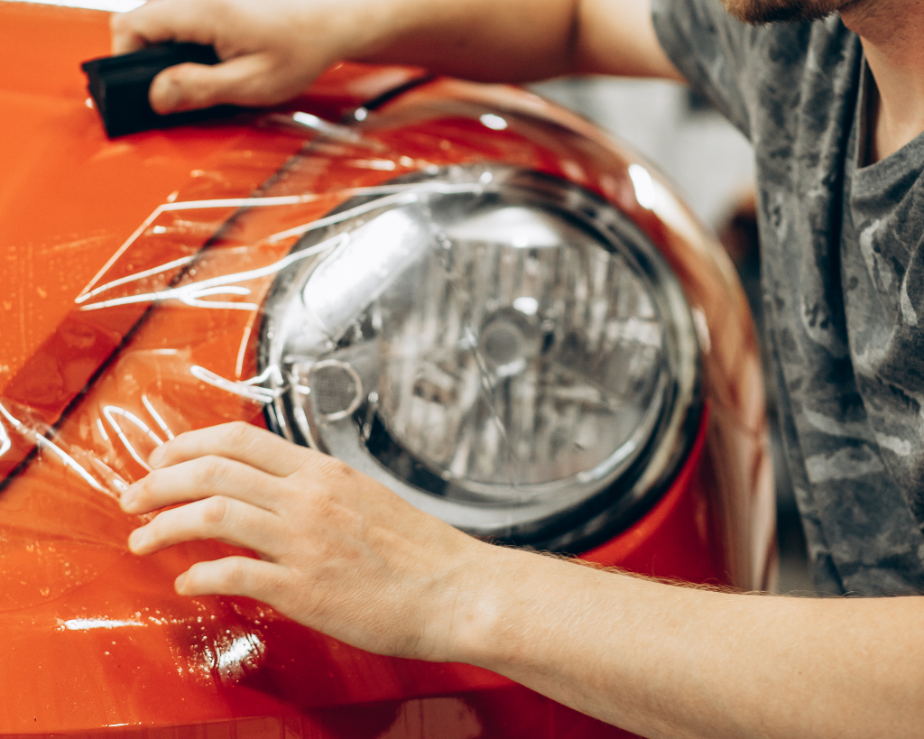 Hands applying clear protective film to an orange car's headlight area.