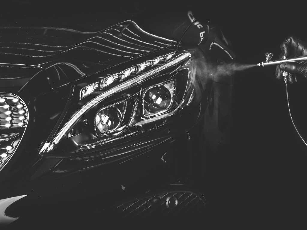 Close-up of a dark car's front end being detailed with a spray gun in a gloved hand, spraying a fine mist near the headlight