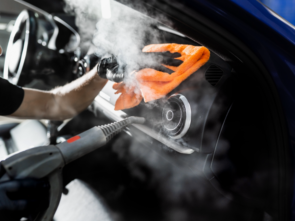 Technician cleaning a car dashboard with a steaming device and an orange microfiber cloth. Close-up of a car interior being steam-cleaned, orange cloth wiping the dashboard. Automotive detailer uses a steam cleaner on a car dashboard, orange microfiber in hand. Steam cleaning a vehicle's dashboard area with a bright orange cloth in use. Dashboard being cleaned with steam and orange microfiber cloth; steering wheel visible. Car detailing: steaming the instrument panel while wiping with orange cloth.