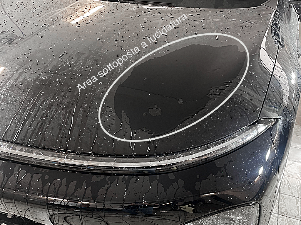 Cofano nero bagnato: fuori dall'area evidenziata l'acqua forma gocce distinte, dentro si stende in un velo continuo perché la zona è stata lucidata e il coating è stato rimosso.